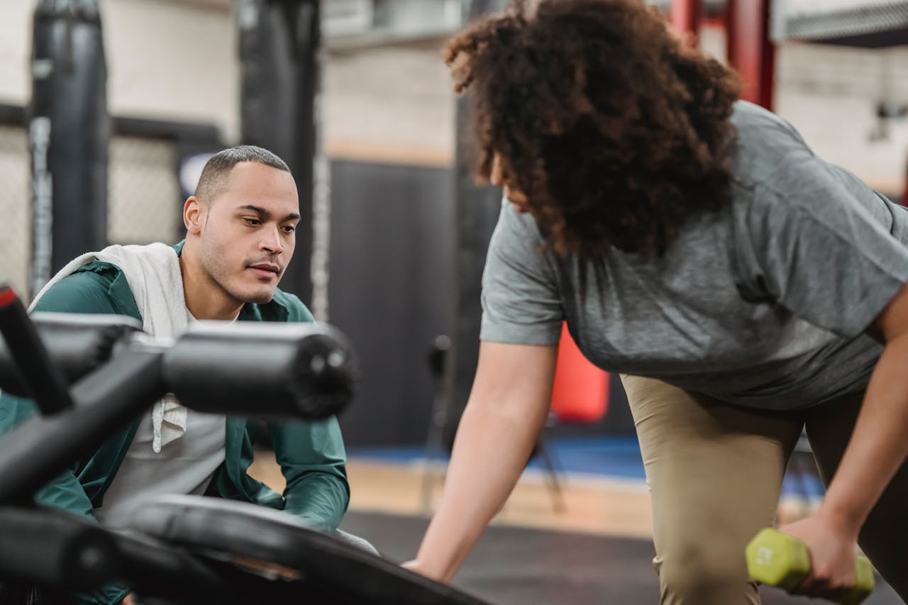 services-04 Experienced instructor watching anonymous African American female lifting dumbbell on blurred background of gym