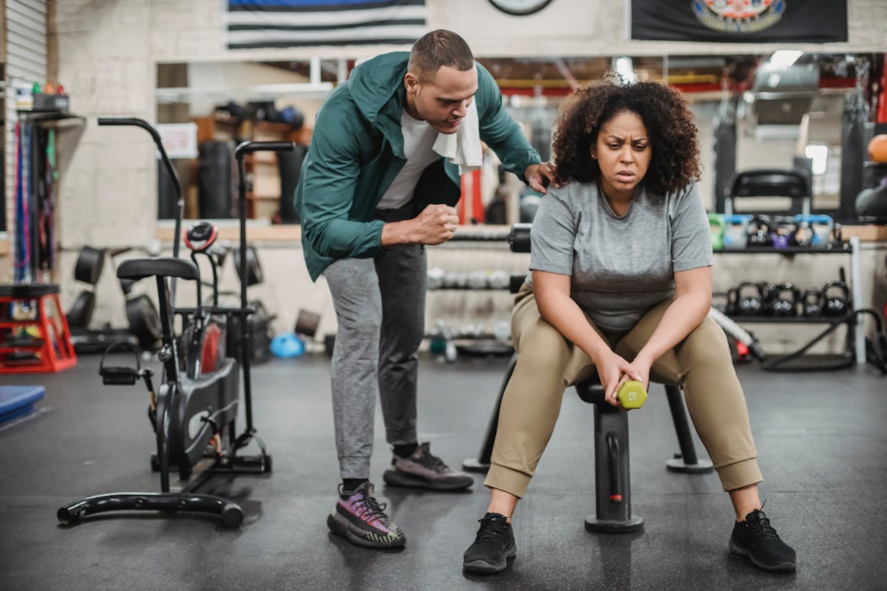 hero-img Full body of male fitness instructor in sportswear supporting tired black woman sitting with dumbbell on bench in gym